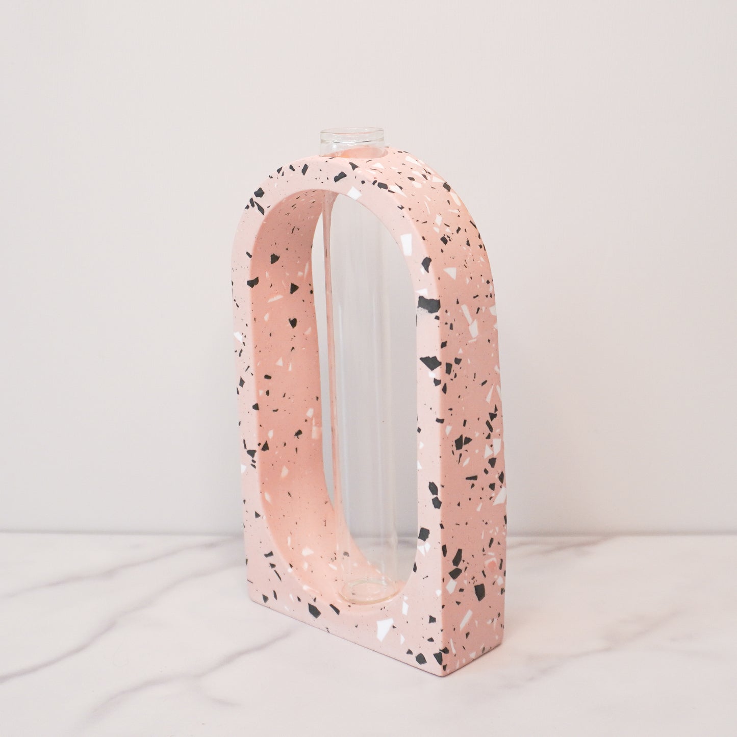 A speckled boho arch propagation vase with a terrazzo-style design in soft pink, featuring white, black, and gray flecks. The arch shape frames a clear glass tube in the center, designed for holding plant cuttings. The vase is displayed on a white marble surface with a light beige background, showcasing its modern and minimalist aesthetic.