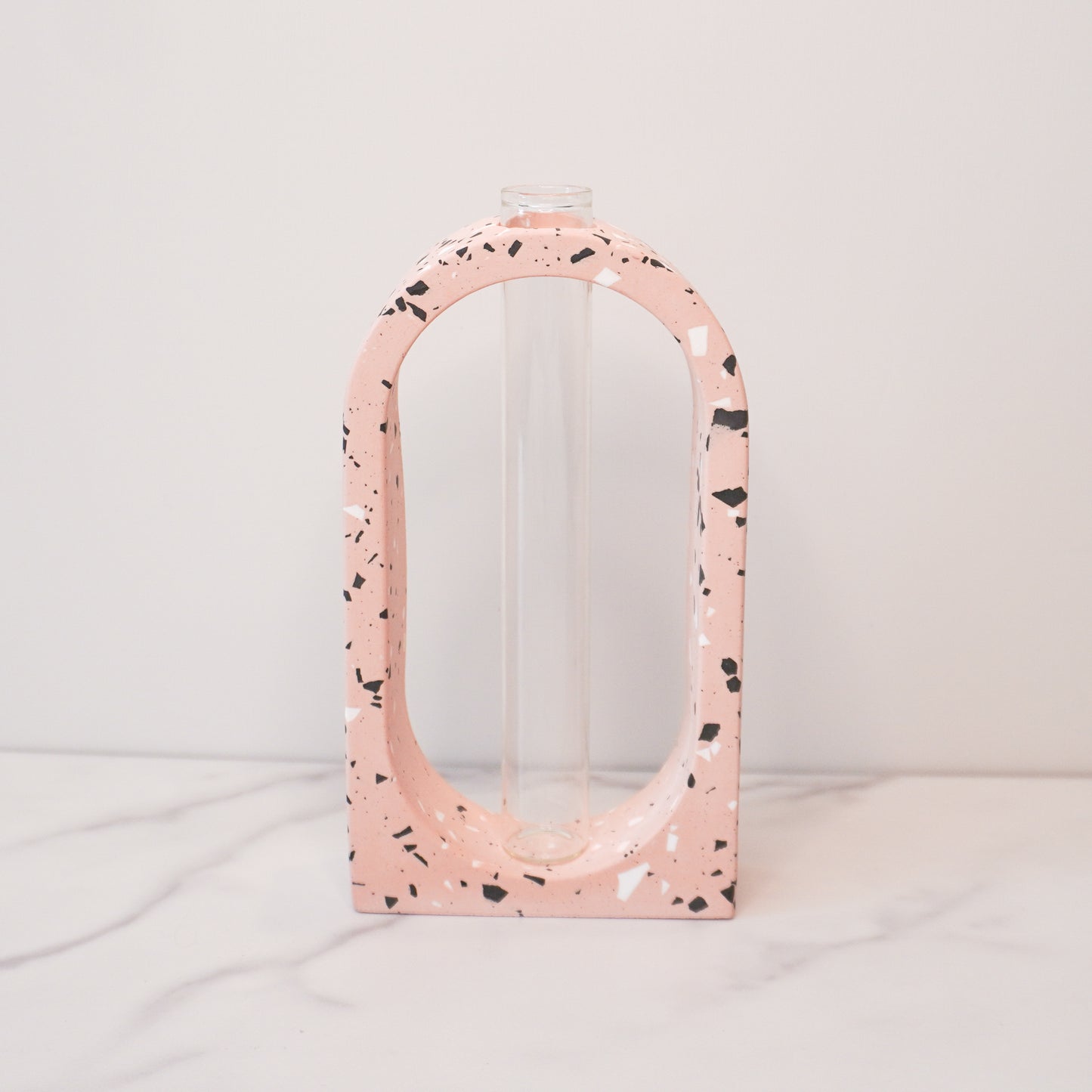 A stylish propagation vase featuring a speckled boho arch design in soft pink with black, white, and gray terrazzo-style flecks. The arch surrounds a clear glass test tube vase in the center, perfect for holding plant cuttings. The vase sits on a white marble surface with a plain white background, highlighting its modern and artistic aesthetic.