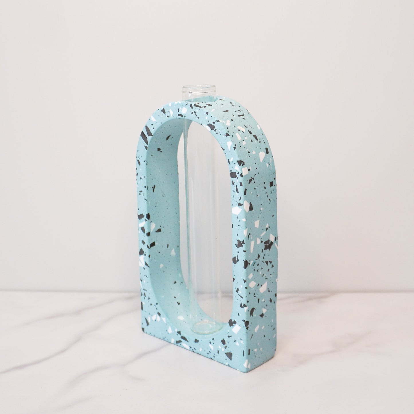 A speckled boho arch propagation vase with a terrazzo-style design in soft blue, featuring white, black, and gray flecks. The arch shape frames a clear glass tube in the center, designed for holding plant cuttings. The vase is displayed on a white marble surface with a light beige background, showcasing its modern and minimalist aesthetic.