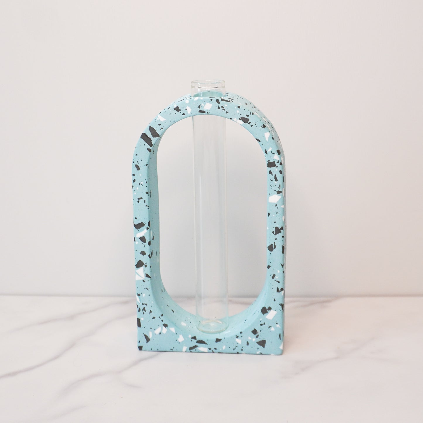A stylish propagation vase featuring a speckled boho arch design in soft blue with black, white, and gray terrazzo-style flecks. The arch surrounds a clear glass test tube vase in the center, perfect for holding plant cuttings. The vase sits on a white marble surface with a plain white background, highlighting its modern and artistic aesthetic.