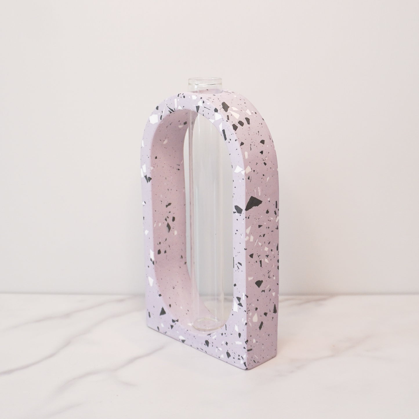 A speckled boho arch propagation vase with a terrazzo-style design in soft purple, featuring white, black, and gray flecks. The arch shape frames a clear glass tube in the center, designed for holding plant cuttings. The vase is displayed on a white marble surface with a light beige background, showcasing its modern and minimalist aesthetic.
