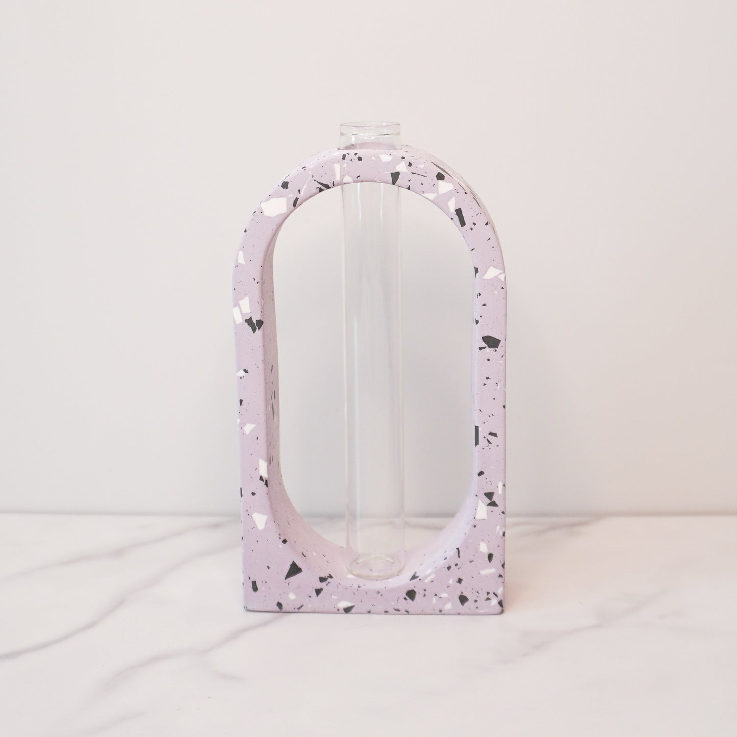A stylish propagation vase featuring a speckled boho arch design in soft lavender with black, white, and gray terrazzo-style flecks. The arch surrounds a clear glass test tube vase in the center, perfect for holding plant cuttings. The vase sits on a white marble surface with a plain white background, highlighting its modern and artistic aesthetic.