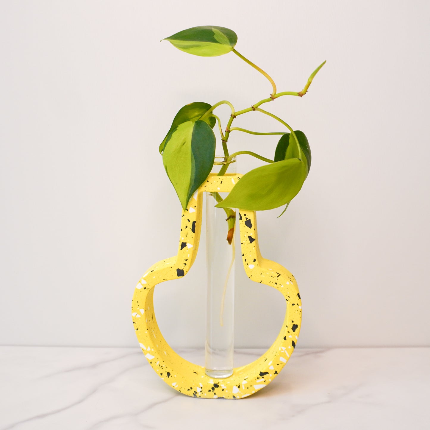 A speckled jar-shaped propagation vase with a modern terrazzo-style design in vibrant yellow, featuring black, white, and gray flecks. The jar-shaped frame surrounds a clear glass test tube vase in the center, holding a green plant cutting with heart-shaped leaves and visible roots. The vase is displayed on a white marble surface with a plain white background, showcasing its bold, artistic, and contemporary design.