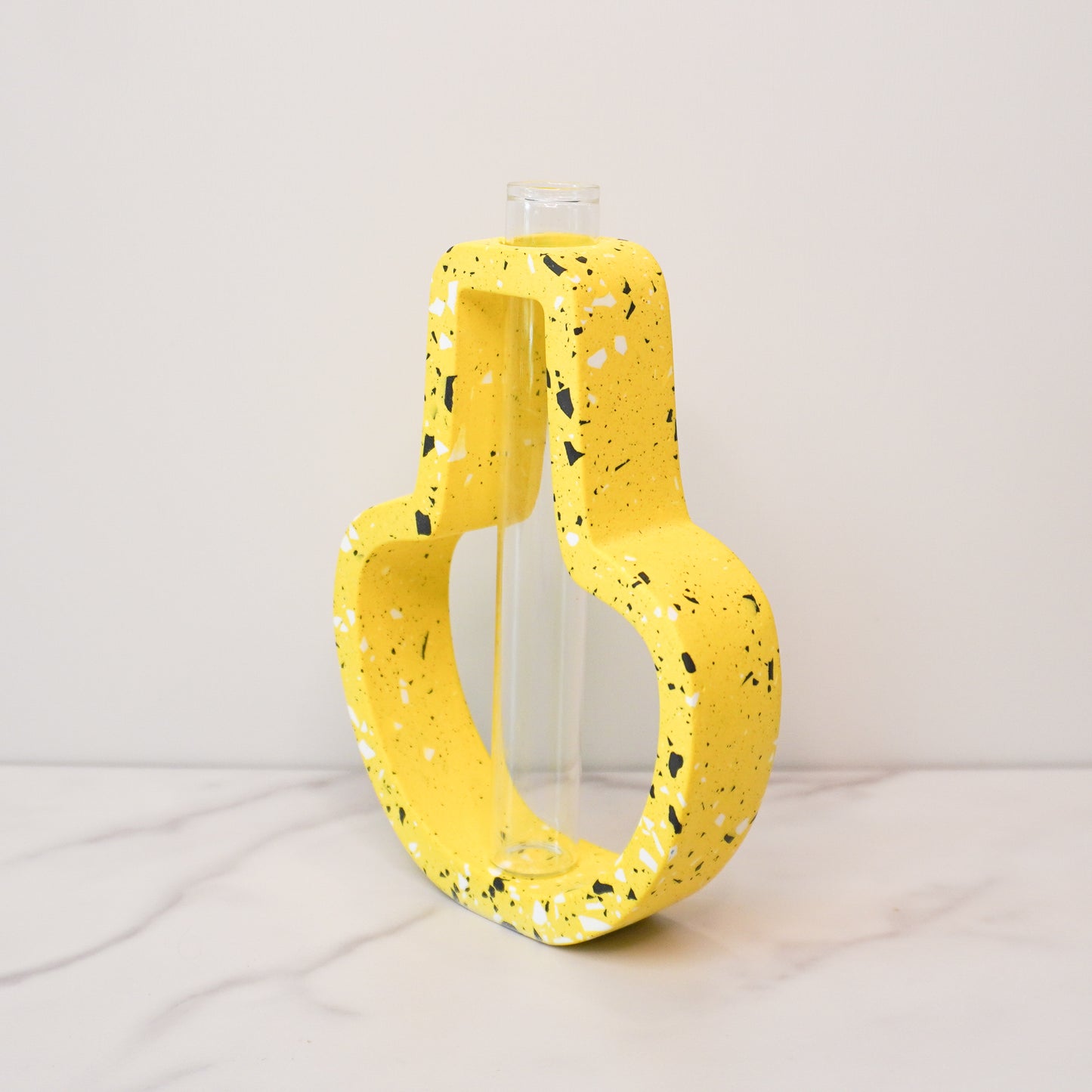 A vibrant propagation vase with a jar-shaped, speckled design in bright yellow, accented with black, white, and gray terrazzo-style flecks. The open-frame structure surrounds a clear glass test tube in the center, designed for holding plant cuttings. The vase is displayed on a white marble surface against a light gray background, showcasing its playful and modern aesthetic.