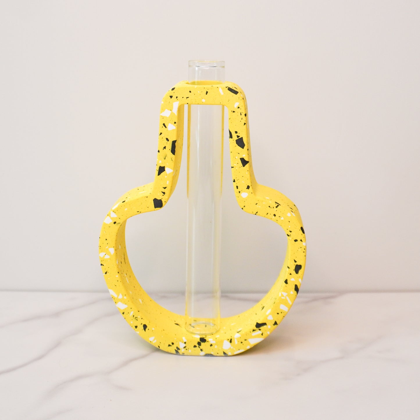 A vibrant propagation vase with a jar-shaped, speckled design in bright yellow, accented with black, white, and gray terrazzo-style flecks. The open-frame structure surrounds a clear glass test tube in the center, designed for holding plant cuttings. The vase is displayed on a white marble surface against a light gray background, showcasing its playful and modern aesthetic.
