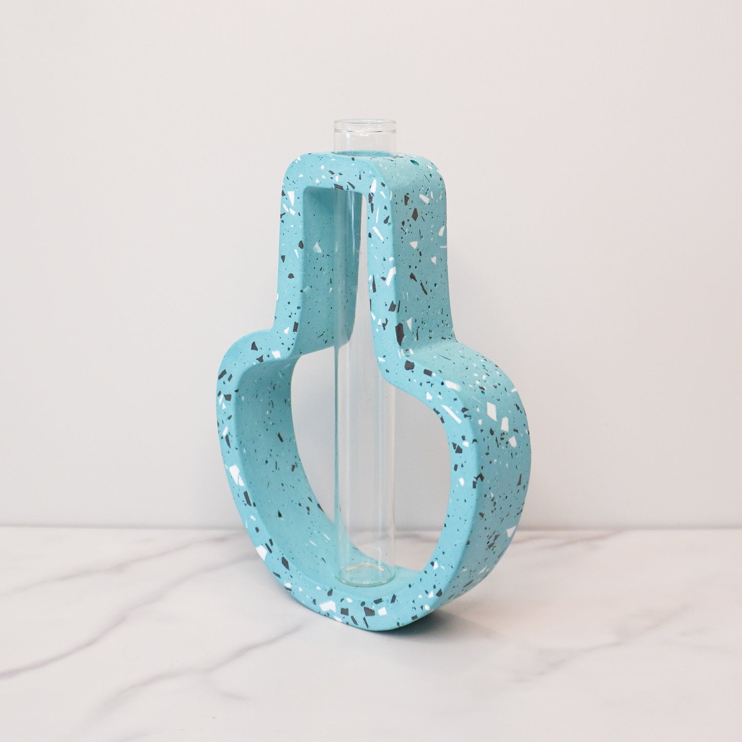 A vibrant propagation vase with a jar-shaped, speckled design in light blue, accented with black, white, and gray terrazzo-style flecks. The open-frame structure surrounds a clear glass test tube in the center, designed for holding plant cuttings. The vase is displayed on a white marble surface against a light gray background, showcasing its playful and modern aesthetic.