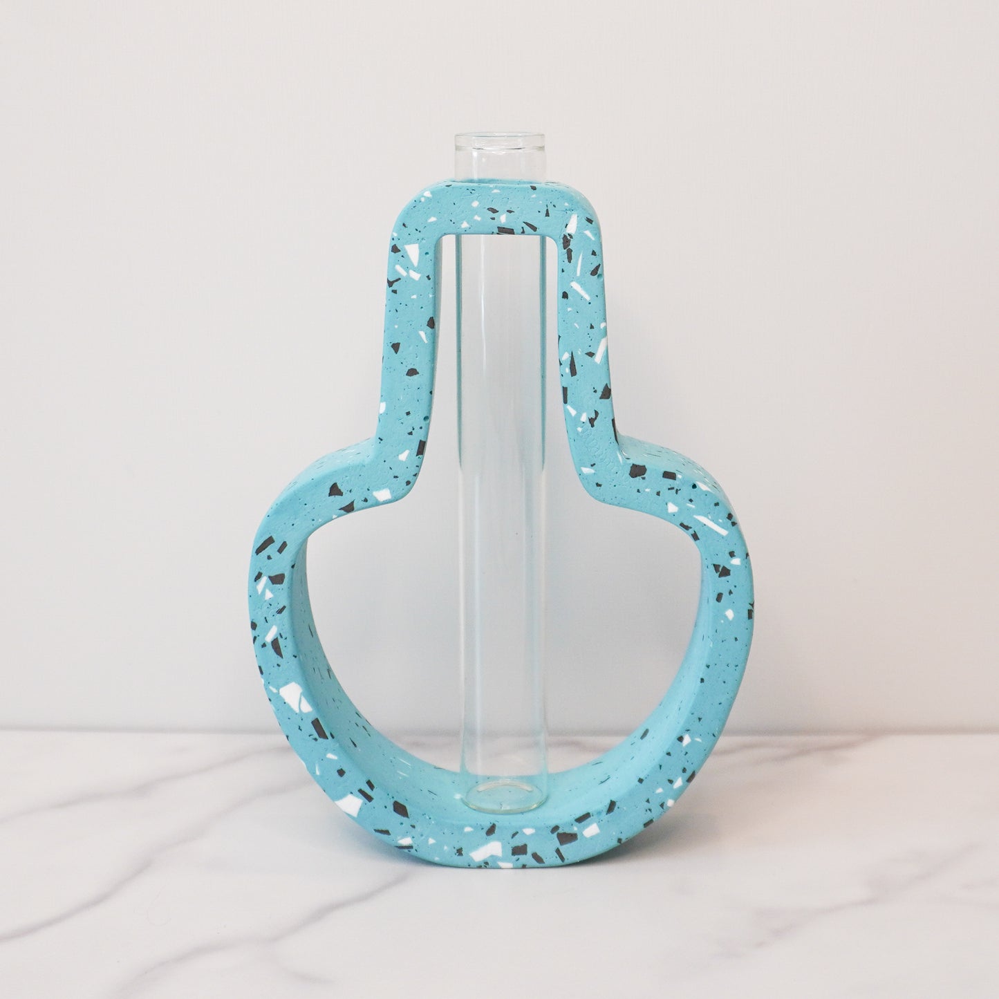 A vibrant propagation vase with a jar-shaped, speckled design in light blue, accented with black, white, and gray terrazzo-style flecks. The open-frame structure surrounds a clear glass test tube in the center, designed for holding plant cuttings. The vase is displayed on a white marble surface against a light gray background, showcasing its playful and modern aesthetic.