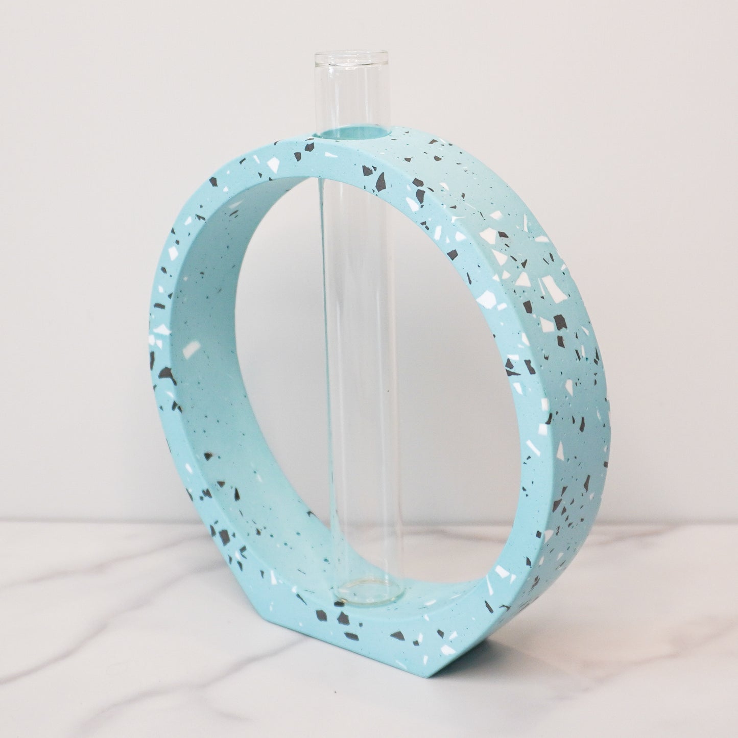 A speckled circle propagation vase with a modern terrazzo-style design in light blue, featuring black, white, and gray flecks. The circular frame surrounds a clear glass test tube vase in the center, designed to hold plant cuttings. The vase sits on a white marble surface with a neutral background, showcasing its artistic and contemporary aesthetic.