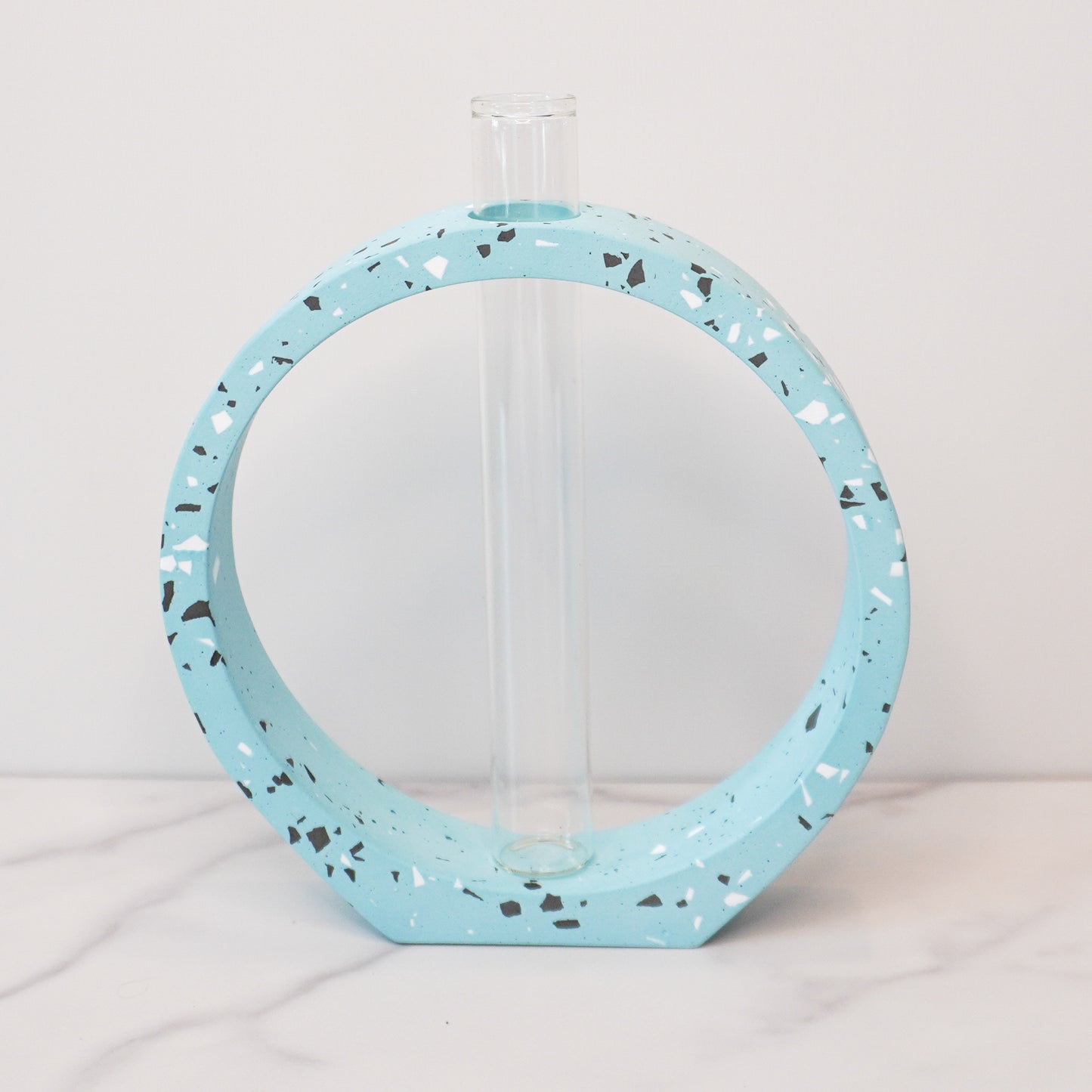 A speckled circle propagation vase with a modern terrazzo-style design in light blue, featuring black, white, and gray flecks. The circular frame surrounds a clear glass test tube vase in the center, designed to hold plant cuttings. The vase sits on a white marble surface with a neutral background, showcasing its artistic and contemporary aesthetic.