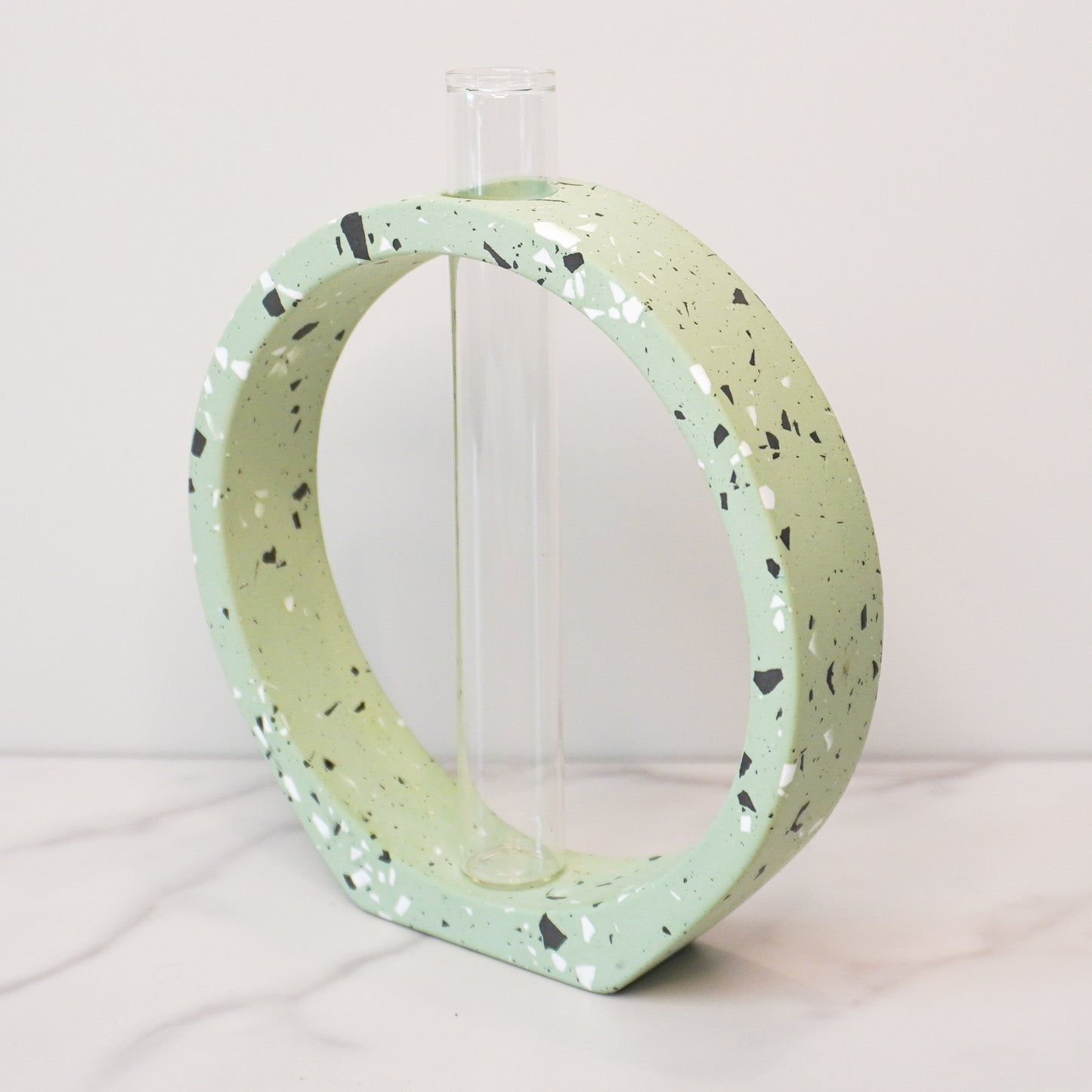 A speckled circle propagation vase with a modern terrazzo-style design in light green, featuring black, white, and gray flecks. The circular frame surrounds a clear glass test tube vase in the center, designed to hold plant cuttings. The vase sits on a white marble surface with a neutral background, showcasing its artistic and contemporary aesthetic.