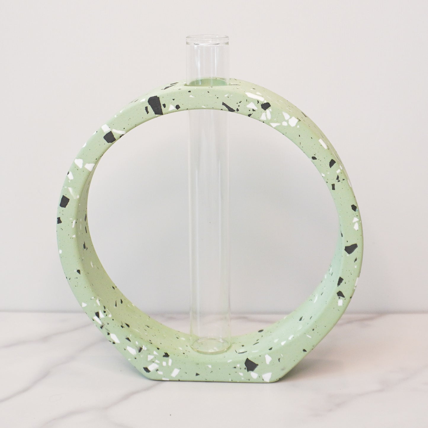 A speckled circle propagation vase with a modern terrazzo-style design in light green, featuring black, white, and gray flecks. The circular frame surrounds a clear glass test tube vase in the center, designed to hold plant cuttings. The vase sits on a white marble surface with a neutral background, showcasing its artistic and contemporary aesthetic.