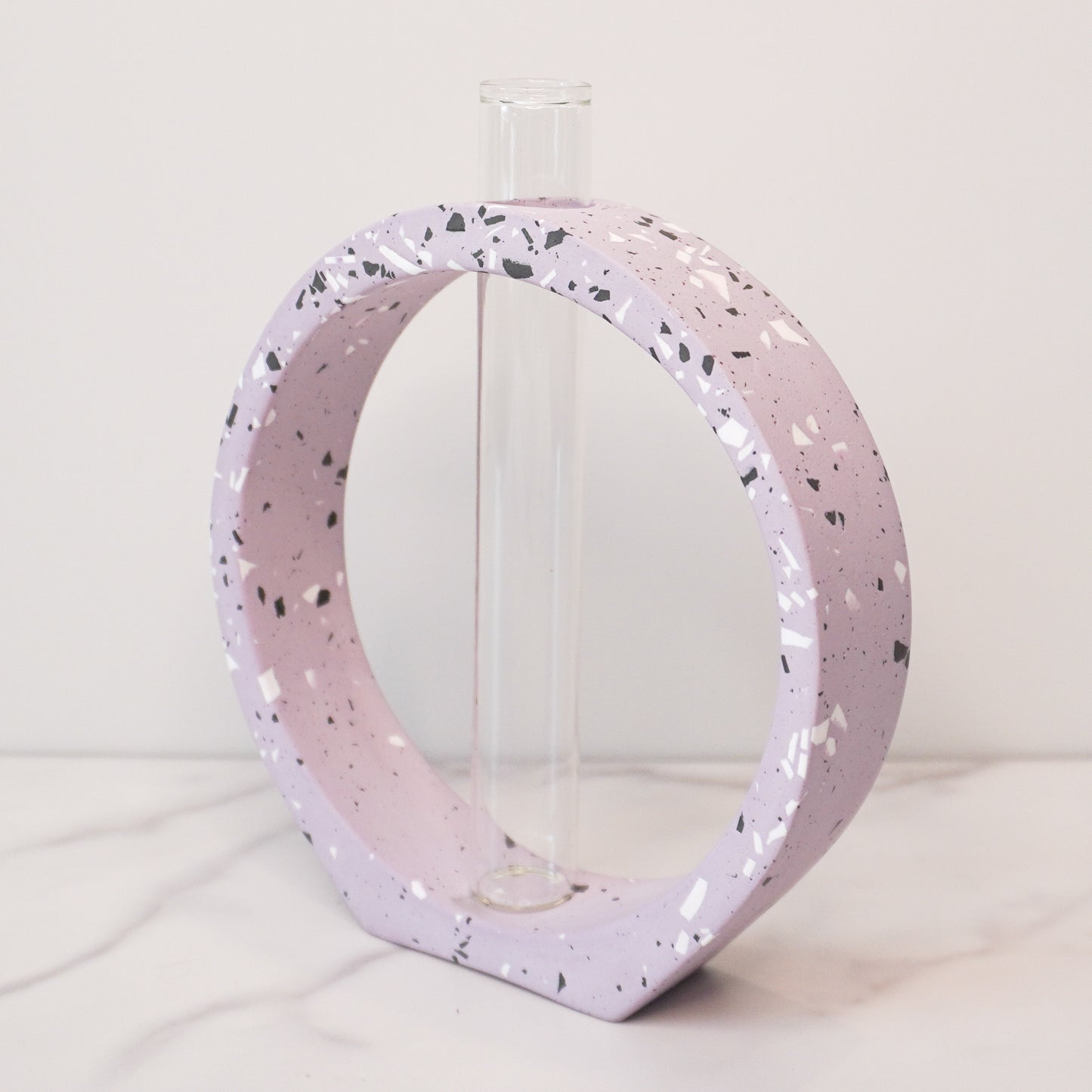 A speckled circle propagation vase with a modern terrazzo-style design in light lavendar, featuring black, white, and gray flecks. The circular frame surrounds a clear glass test tube vase in the center, designed to hold plant cuttings. The vase sits on a white marble surface with a neutral background, showcasing its artistic and contemporary aesthetic.
