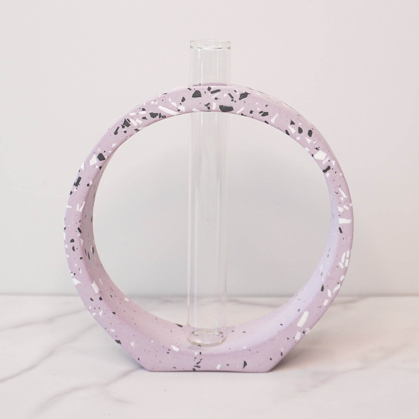 A speckled circle propagation vase with a modern terrazzo-style design in light lavendar, featuring black, white, and gray flecks. The circular frame surrounds a clear glass test tube vase in the center, designed to hold plant cuttings. The vase sits on a white marble surface with a neutral background, showcasing its artistic and contemporary aesthetic.