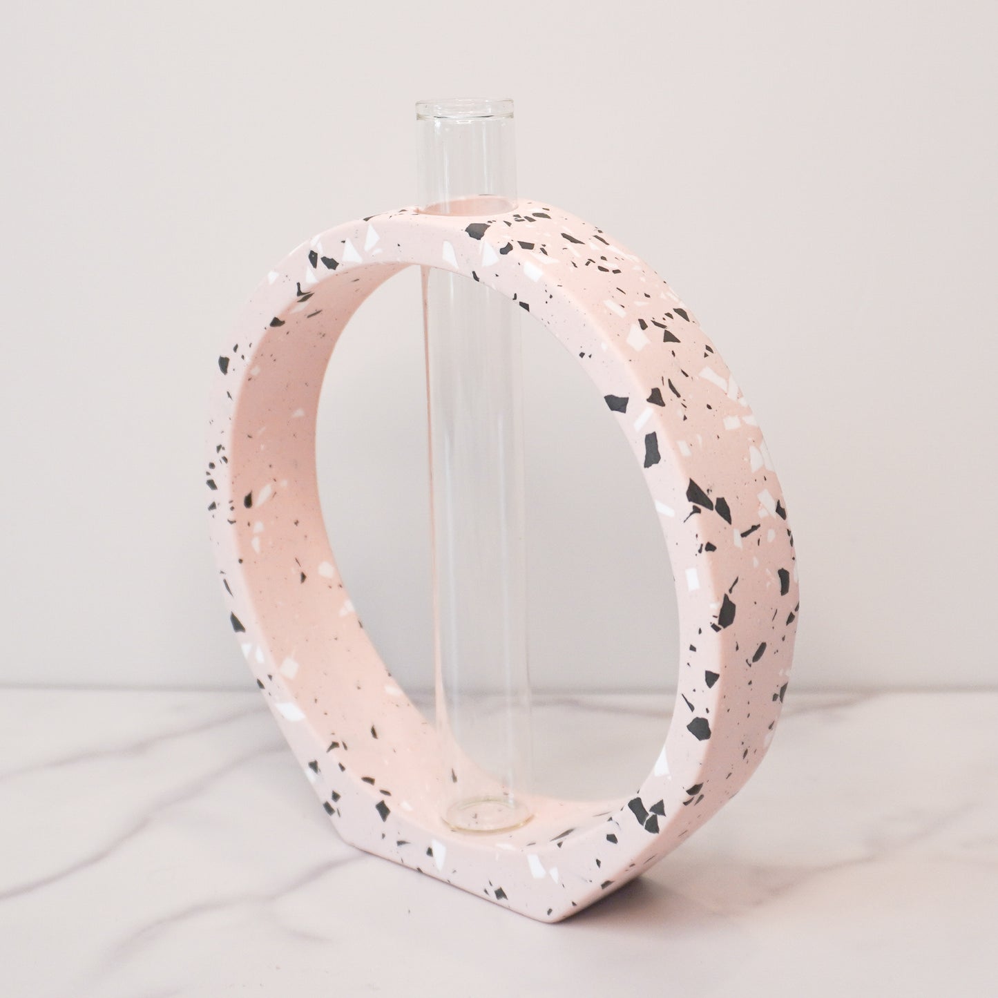 A speckled circle propagation vase with a modern terrazzo-style design in light pink, featuring black, white, and gray flecks. The circular frame surrounds a clear glass test tube vase in the center, designed to hold plant cuttings. The vase sits on a white marble surface with a neutral background, showcasing its artistic and contemporary aesthetic.