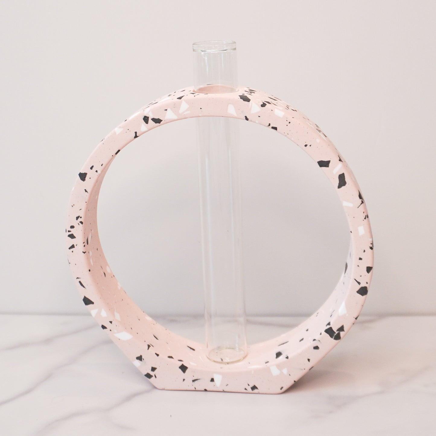 A speckled circle propagation vase with a modern terrazzo-style design in light pink, featuring black, white, and gray flecks. The circular frame surrounds a clear glass test tube vase in the center, designed to hold plant cuttings. The vase sits on a white marble surface with a neutral background, showcasing its artistic and contemporary aesthetic.
