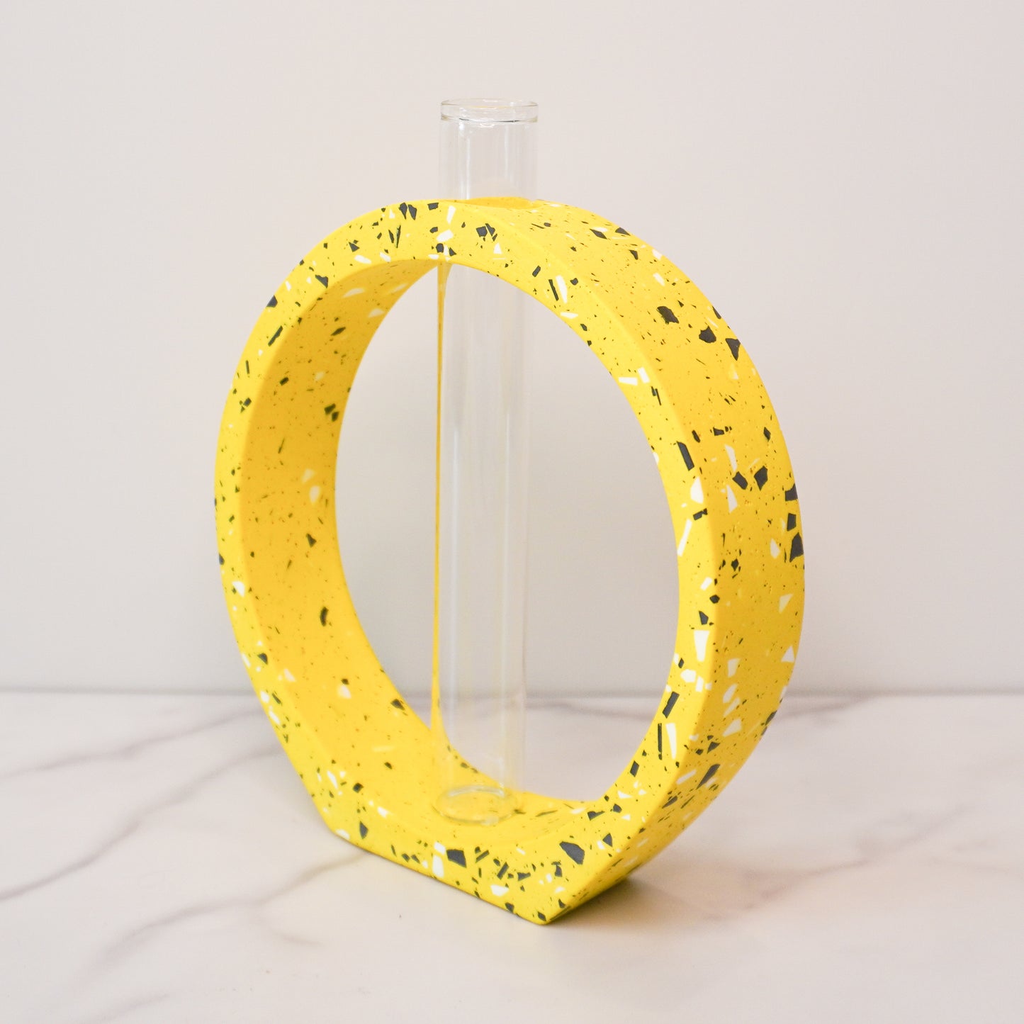 A speckled circle propagation vase with a modern terrazzo-style design in bright yellow, featuring black, white, and gray flecks. The circular frame surrounds a clear glass test tube vase in the center, designed to hold plant cuttings. The vase sits on a white marble surface with a neutral background, showcasing its artistic and contemporary aesthetic.