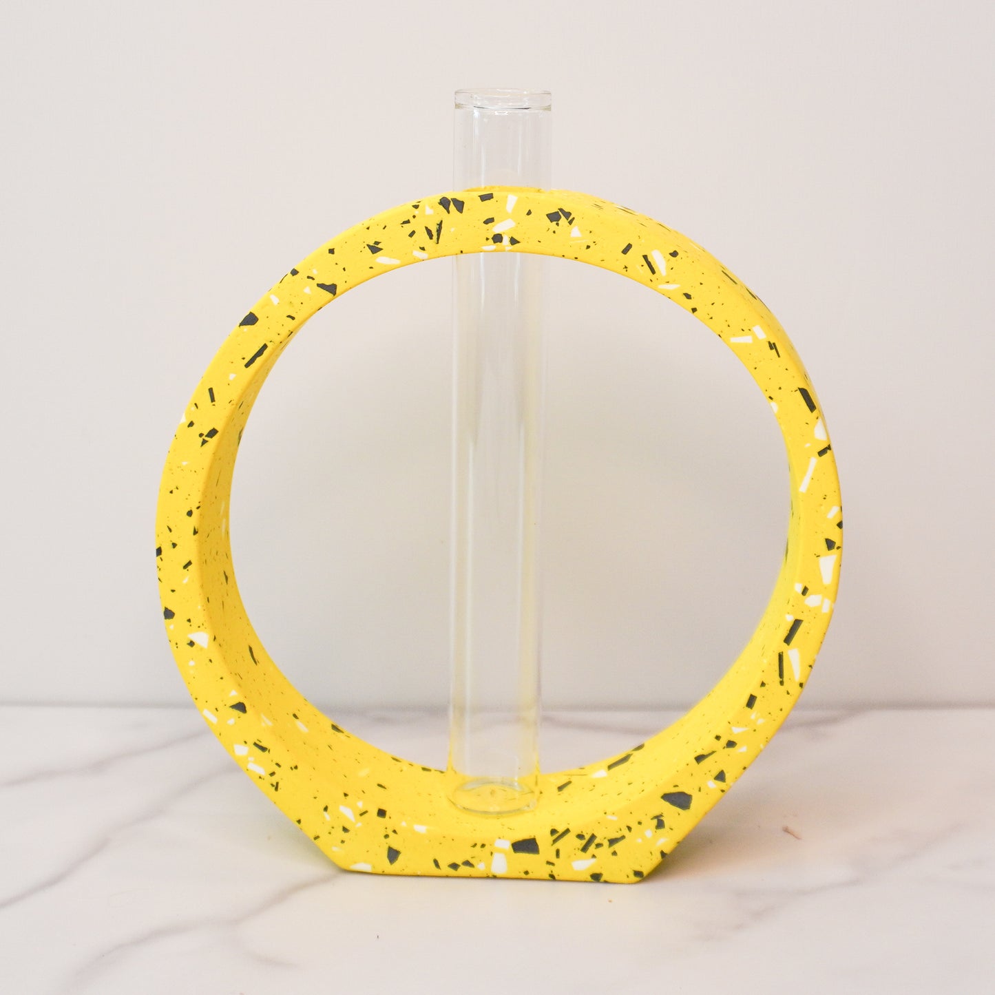 A speckled circle propagation vase with a modern terrazzo-style design in bright yellow, featuring black, white, and gray flecks. The circular frame surrounds a clear glass test tube vase in the center, designed to hold plant cuttings. The vase sits on a white marble surface with a neutral background, showcasing its artistic and contemporary aesthetic.