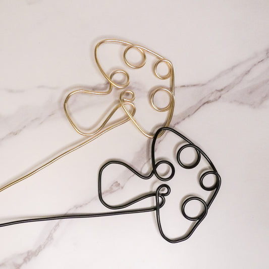 Two mushroom-shaped wire plant trellises displayed on a white marble surface. One trellis is crafted in a shiny gold finish, while the other features a sleek black finish. Both designs incorporate whimsical circular cutouts, adding a playful and functional touch for supporting climbing plants.