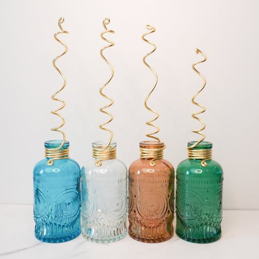 Four vintage-style glass bottles in vibrant colors: blue, clear, amber, and green. Each bottle features intricate embossed patterns and is topped with a whimsical gold spiral wire trellis extending upward. The bottles are arranged in a row on a white surface against a plain white background, highlighting their decorative and functional design.