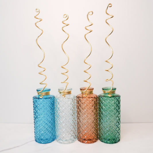 Four quilted glass bottles in vibrant colors—blue, amber, green, and clear—are arranged side by side, each topped with a gold spiral wire trellis. The bottles feature a textured diamond pattern, adding a decorative touch, while the trellises extend upward in whimsical spirals, designed to support climbing plants. The setup is displayed against a plain white background, highlighting the playful yet elegant design.