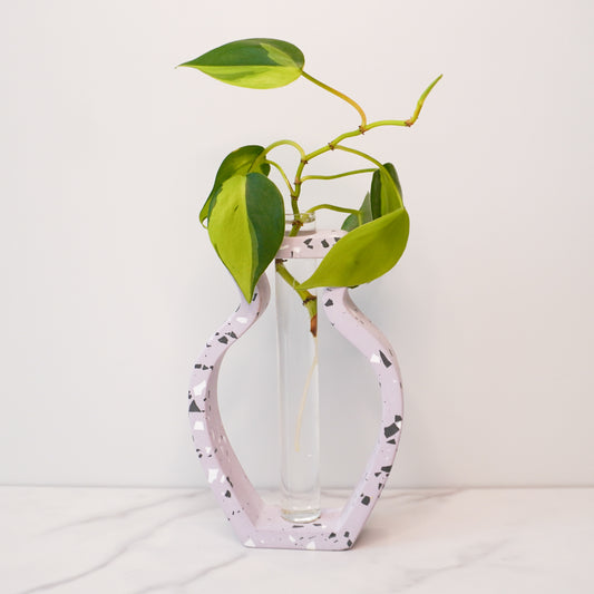 A speckled vase-shaped propagation vase with a terrazzo-style design in soft lavender, featuring black, white, and gray flecks. The open-frame structure holds a clear glass test tube in the center, containing a vibrant green plant cutting with heart-shaped leaves and visible roots. The vase is displayed on a white marble surface against a plain white background, emphasizing its modern and artistic design.