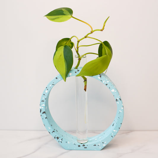 A modern propagation vase featuring a speckled circular design in light blue with white, black, and gray terrazzo-style flecks. The circle frames a clear glass test tube in the center, holding a green plant cutting with heart-shaped leaves and visible roots. The vase is displayed on a white marble surface against a plain white background, highlighting its artistic and minimalist aesthetic.