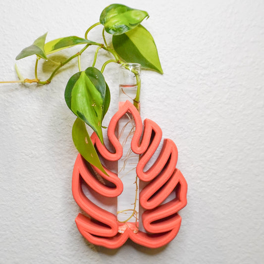 A wall-mounted Monstera-inspired propagation tube holder made of rust colored material with a cutout leaf design. A clear glass tube is inserted in the center, filled with water and holding green pothos cuttings with visible roots. The holder is displayed against a white textured wall, with the vibrant leaves adding a natural touch.