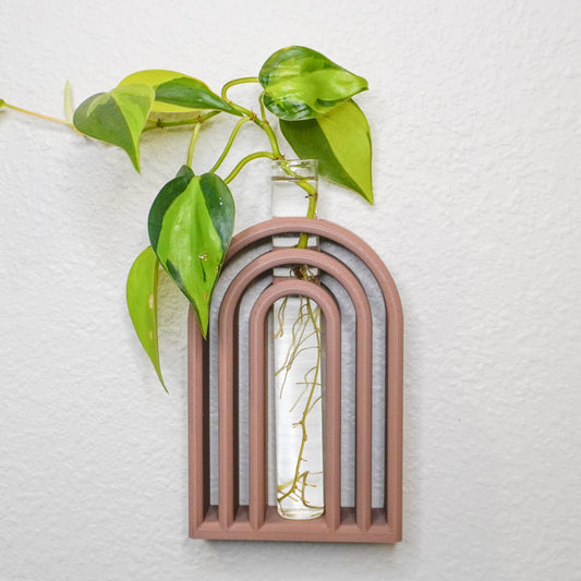 A Boho-style wall-mounted propagation tube holder shaped like an arched frame with layered details in a warm terracotta color. A clear glass tube is centered within the arch, holding water and vibrant pothos plant cuttings with green and yellow variegated leaves. The piece is displayed on a textured white wall.